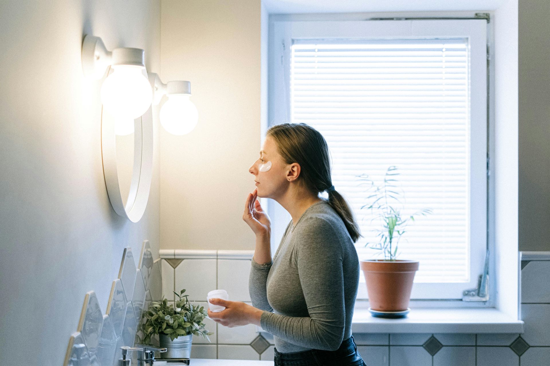 Kobieta pielęgnuje twarz przed oświetlonym lustrem w łazience  https://www.pexels.com/pl-pl/zdjecie/kobieta-lazienka-trzymajacy-krem-4491161/